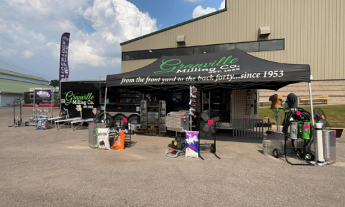 Outdoor setup of Granville Milling Co. show trailer featuring a branded canopy with the slogan 'from the front yard to the back forty...since 1953.' The display includes various outdoor equipment, tools, and promotional items in front of a large building.