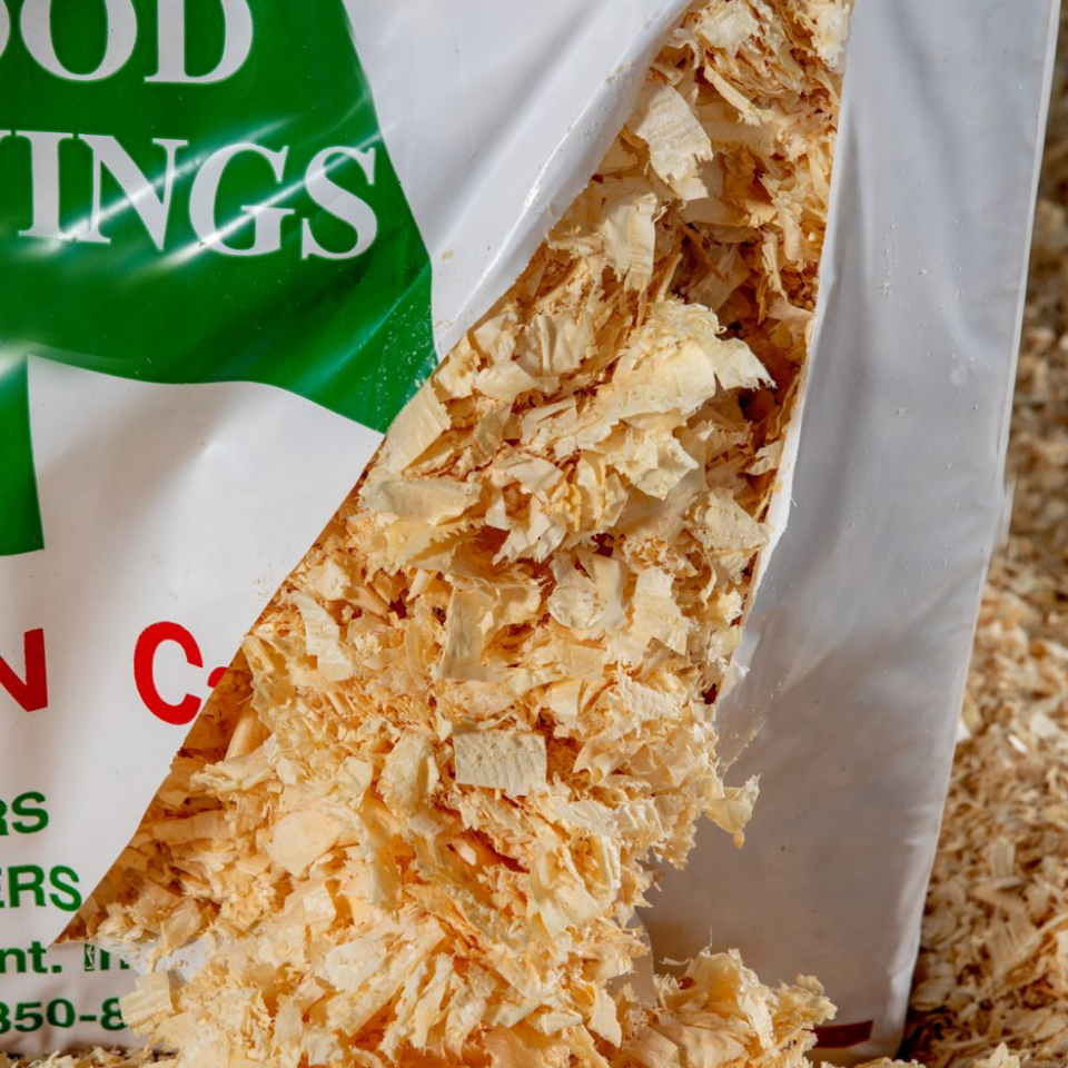 Yellow bag of wood shavings labeled “McFEETERS Ultimate Quick Pick Bedding” with green text and tree graphic, surrounded by loose shavings.